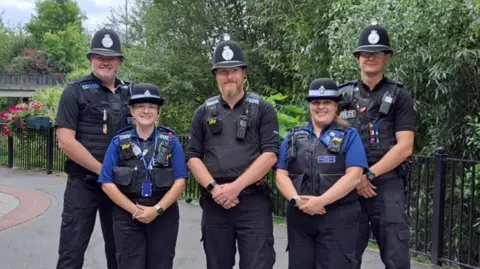 West Mercia Police A group of five police officers, three men and two women, stand together in a pedestrianised area smiling at the camera.