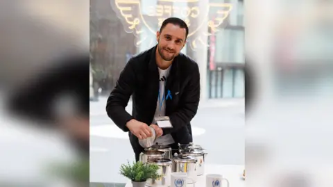 UK for UNHCR Smiling Mouhyedin Alkhalil pouring tea into silver pots on a table with a green plant in the foreground
