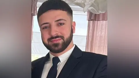 Hertfordshire Police Kyle Clifford with dark hair and beard wearing a black jacket, white shirt and black tie