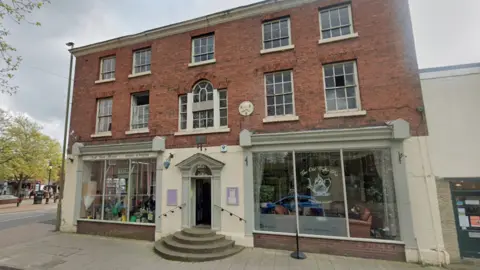 Google A large building housing two shops. The top half is red brick and the bottom half has large windows wither side of a door. There are stone steps leading to the door, which is green and cream and has black hand rails.