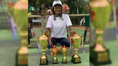 Eddy Edwards Marylove Edwards kneels in front of tennis net with four golden trophies