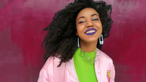 Christina Mallas Yesenia smiles in front of a pink wall, wearing a green jumper and a pink jacket