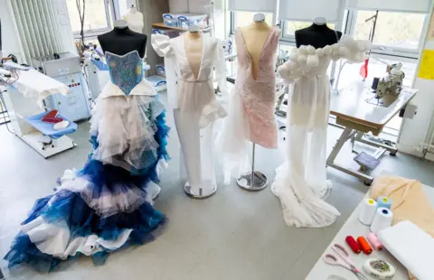 StoryShop Four dress designs are tacked onto mannequins in a work shop. The first one has a blue sparkly bodice with a blue and white mesh skirt. The second is white and has a deep V neck and asymmetric detailing in the shoulders and skirt. The second is light pink and about knee length with a very deep V down to about the hips. The fourth has white balls about the size of a fist going diagonally across the dress. It has a short skirt with a longer mesh skirt over the top.