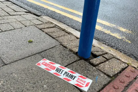 Photo of pavement with 'CAUTION WET PAINT' sticker surrounded by blue paint