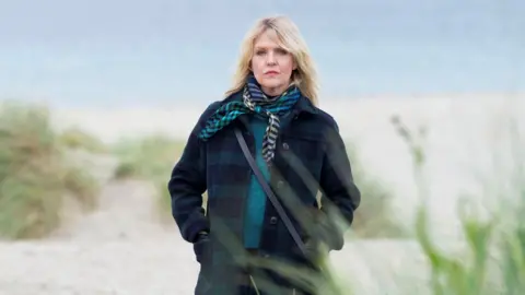 BBC/Silverprint Films/Jamie Simpson Ashley Jensen stands on a deserted Shetland beach, the sea grey behind her. She is wrapped up in a thick green and navy tartan wool coat with a teal woolly jumper and a patterned scarf. Her hands are in her pockets as she looks moodily at the camera.