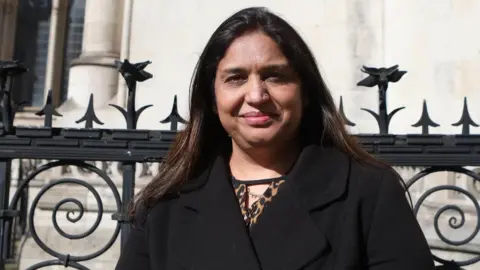 PA Media Seema Misra standing in front of some black railings, looking directly at the camera with a half smile. She is wearing a black coat. 