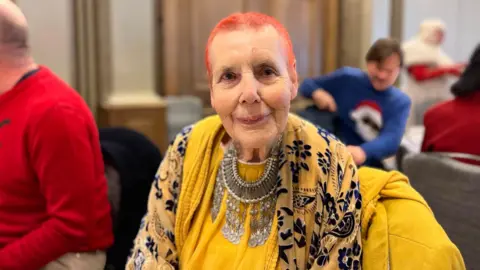 George Carden/BBC Hilary Howard at the festive party. She is wearing a bright yellow top, yellow jacket and a large necklace. She has orange hair and is looking directly at the camera.