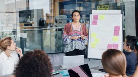 Getty Images A Spanish female is heading up a business meeting to female colleagues in a glass cubicle office