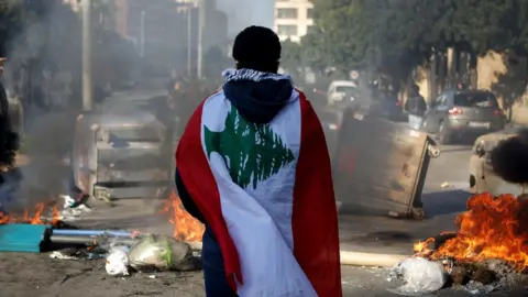 AFP A Lebanese anti-government protester, wrapped in a national flag, stands in front of a road blocked with burning tyres and overturned rubbish bins (14 January 2020)