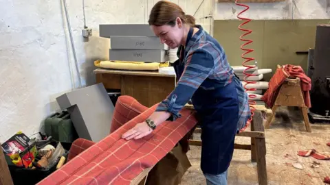 BBC A woman with tied-back brown hair works on reupholstering a sofa lying on its back, stretching patterned red fabric over the frame, surrounded by tools in a workshop.