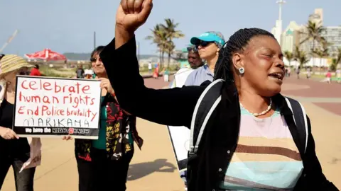 AFP A protester raises her fist as others hold up a banner reading 'Celebrate human rights, End police brutality' during the commemoration of the third anniversary of the Marikana massacre, at the north beach in Durban, on August 16, 2015.
