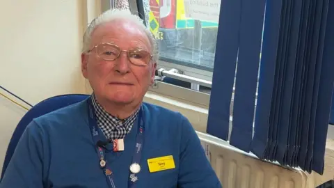 BBC Terry Seston, who has grey hair and gold glasses is wearing a checked shirt and blue jumper, sits in front of a window. Behind him, out of the window, can be seen a yellow and blue ambulance vehicle.