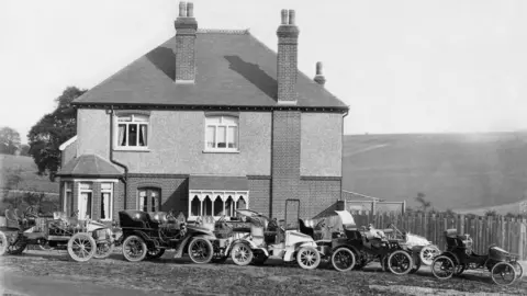 Stellantis A black and white photo of a detached house with carriage style motor cars parked outside. There are hills in the background.
