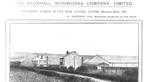 Stellantis A black and white photo of an industrial looking shed stands in fields and next to some houses.