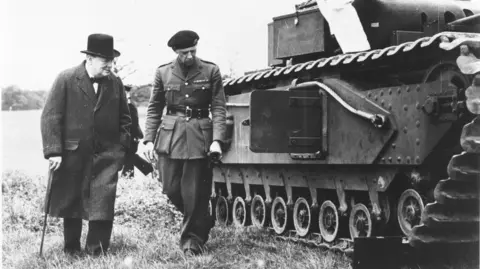 Vauxhall Heritage Winston Churchill, holding a walking stick and wearing a hat and heavy coat, stands next to a person in uniform while they book look at a tank in a field.