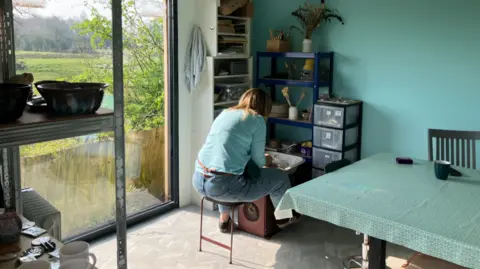 A woman can keep be seen leaning over a potters wheel, working with clay. To the side of her is a large window looking out onto a field.