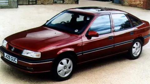PA Media A burgundy saloon-style car, with a L-reg number plate, stands on a gravel driverway. 