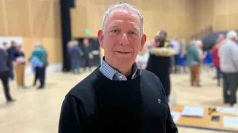 BBC A man with grey hair in a dark blue jumper with a blue and white striped shirt underneath