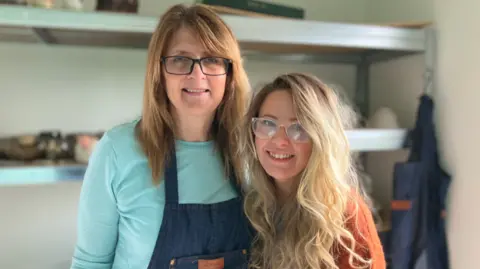 BBC Two women, both wearing glasses, stand close together and are smiling. The woman on the left has long brown hair and is wearing dark blue dungarees over a light blue top, while the woman on the right, who has long blond hair, is wearing an orange woollen jumper.
