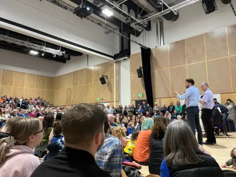 A man in a blue shirt talks to a hall packed with people gathered round to listen