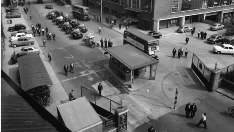 Stellantis A black and white photo of gates to the factory plant with a check-in area, and buses and vehicles dotted around.
