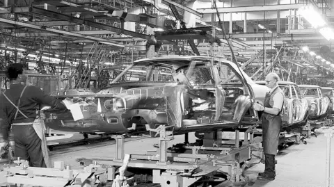 Stellantis A black and white photo of an elderly man wearing spectacles and an apron, with his shirt sleeves rolled up stands on the car production line holding a hose.
