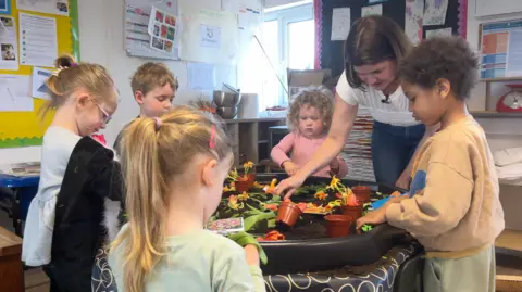A brunette woman leads a group of small children in a table game at a nursery in Somerset.