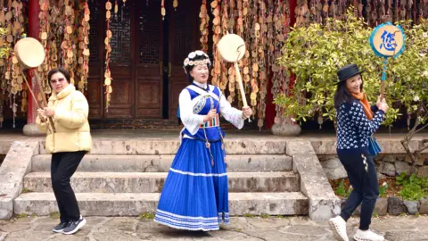 BBC/Rachel Yu Two travellers either side of a traditional Naxi dancer wearing a blue and white dress and petal-like ornamental headgear - all holding a long-handled drum - keep time to music with steps leading to the door of a cultural centre.