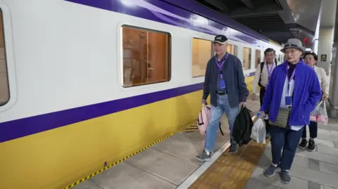 BBC/Benjamin Begley A group of four people - two men and two women - carrying bags in their hands, the front two wearing hats and blue tops, walk along the platform of a train with stripes of blue, white and yellow