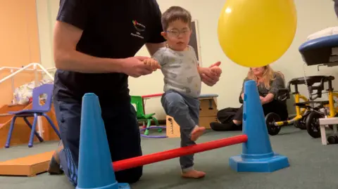 A young child is helped over an obstacle, which is a red stick between two blue traffic-style cones. The child is wearing glasses and being helped by an adult in dark clothing whose head is not visible.
