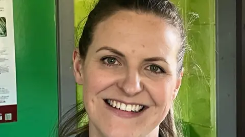 Ffion, a woman with brown hair tied back, smiling in front of a green wall