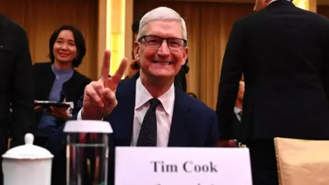 Getty Images Apple's CEO Tim Cook, in a navy blue suit and striped tie, smiles as he holds his fingers up in a victory sign at a meeting in Beijing last year. 