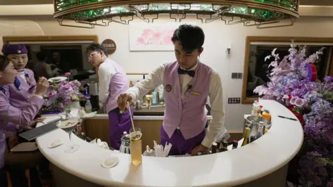 BBC/Benjamin Begley A bartender wearing a pink vest, purple trousers and black bowtie pouring a drink on to a glass sitting on top of a white semi-circular counter.