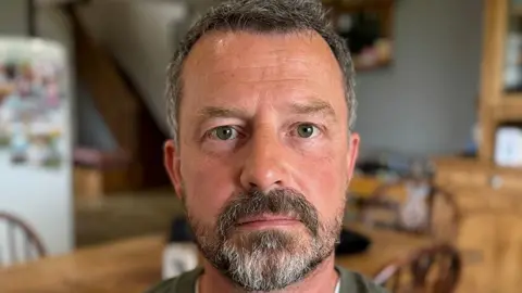 BBC / Sarah Sanderson Simon Skelton looking at the camera. He has dark beard and hair and is standing in a kitchen