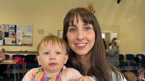 BBC Katie, with long brown hair and a fringe, is smiling and holding a baby wearing a pink cardigan and striped dress holding a toy