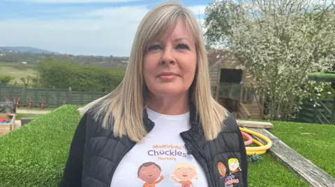 Lisa, with shoulder-length straight blonde hair, wearing a t-shirt and black jacket which says Meithrinfa Chuckles Nursery with a graphic of two young children on. 