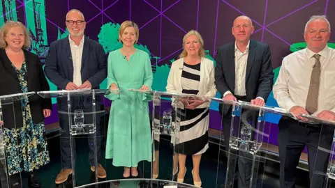 BBC Three men and three women stand behind glass podiums in a TV studio in front of a green and purple backdrop. Sally Horscroft, on the left, wears a floral dress and dark jacket. Jason Stockwood, next left, wears a dark jacket and open-neck white shirt. Dame Andrea Jenkyns wears a light green dress. Marianne Overton wears a black and white dress and cream jacket. Rob Waltham wears a dark suite and open-neck white shirt. Trevor Young wears a white shirt and striped tie.