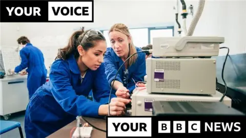 Getty Images Two women wearing blue boiler suits lean towards a machine and work with cables