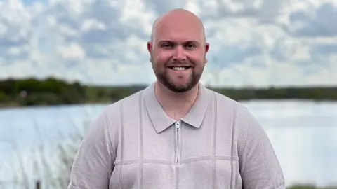 BBC / Sarah Sanderson Harry Finnigan who has a bald head and dark beard he is wearing a grey zipped top and is standing in front of a lake