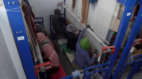 Two rows of three young women, sat back to back, each facing tall looms.