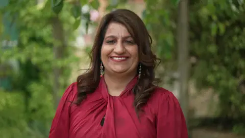 Nishi Gill in a red blouse. She has colourful earrings and brown curly hair. 
