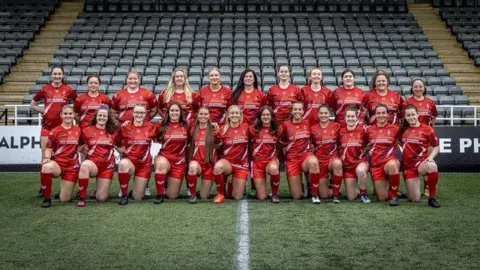 Francesca Johnson-Harding The North East Women's XV squad picture. The 23 players are arranged in two rows, standing at the back and down on one knee in the front. Their kit is all red and they are on a pitch in front of a stand with dark grey seats.