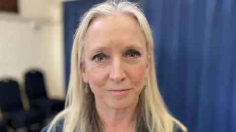 Head and shoulders photo of Dr Roz Savage. She is smiling as she looks at the camera. She has shoulder-length, light blond hair which she is wearing down. She has dark eyeshadow on and is standing in front of a blue curtain and some chairs.