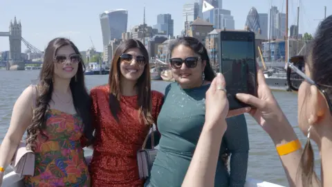 Lovina, centre, posing for a photo with two other women, while standing on a boat on the River Thames in London. All three women are wearing sunglasses, with the lady on the left in a floral dress, Lovina in a red sequin dress and the lady on the right in a green long-sleeved dress.