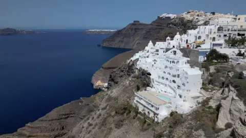 Kevin Church/BBC A photograph showing the cliffs of Santorini with white houses and domes perched on top with green vegetation and blue sea water underneath the cliffs on the volcano's crater rim