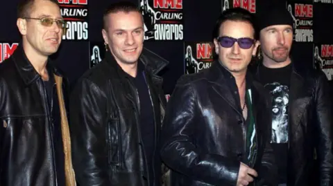 PA Media Adam Clayton, Larry Mullen, Bono and The Edge from U2 in a photograph from 2001. They are standing in front of a sponsorship backdrop for the NME awards.