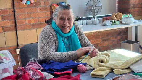 Yorkshire Women's Forum A woman, possibly in her 60s or early 70s, sits with a broad smile behind table which is full of multicoloured bits of material. She is holding some knitting needles and has a ball of wool in front of her.