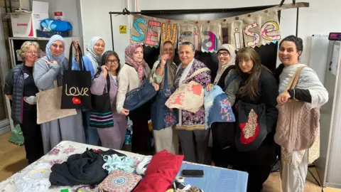 Aisha Iqbal/BBC A group of ladies stand in front of a big sign which says 'Sewcialise'. The sign is made of handmade textile bunting, and the women are all holding up tote bags made of different materials. In front of them is a table full of rolls and piles of colourful textiles, boxes of thread and other related items.