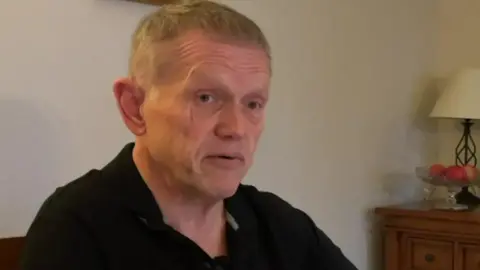 Richard Barker, who has short fair hair and is wearing a black polo shirt, sits in his living room, speaking to a BBC reporter who is out of shot