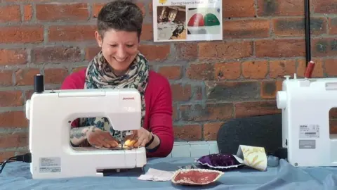 Yorkshire Women's Forum A smiling woman sitting working on a sewing machine. She wears a pink top and a cravat style scarf. She has short, dark pixie style hair.